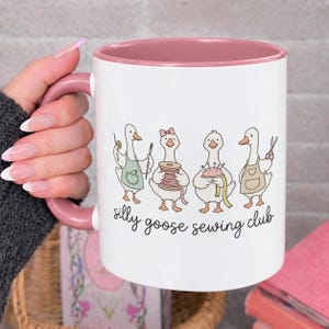 May include: A white ceramic mug with a dusty rose pink handle and interior rim. The mug features a graphic of four cartoon geese engaged in sewing activities, with the text "silly goose sewing club" printed in black script underneath the geese.