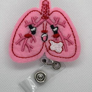 May include: Pink felt embroidered badge reel with a cartoon lung design. The lung has a smiling face with red cheeks and a white bandage on its side. The reel has a clear plastic strap and a silver clip.