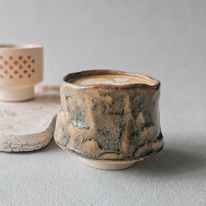 May include: A ceramic cup with a mottled brown and green glaze featuring an uneven rim, positioned next to a smaller, light-colored ceramic cup with a repeating diamond pattern, both resting on a textured, irregular, off-white ceramic tray.