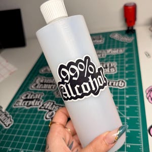 May include: A clear plastic bottle with a white cap, labeled with a black and white sticker that reads "99% Alcohol". The bottle is held in a hand with long, decorated nails. The background includes a green cutting mat and other stickers.