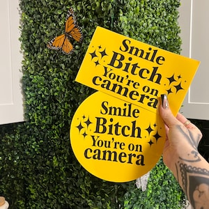 May include: Two yellow signs with black text and glitter accents that read "Smile Bitch You're on camera". One sign is rectangular and the other is round.