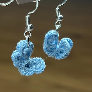 May include: A pair of light blue crochet butterfly earrings featuring silver-toned metal fishhook ear wires. Each earring is crafted from blue yarn in a small, textured butterfly shape, suspended from a silver-colored metal hook for pierced ears.