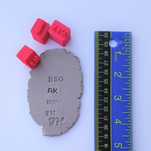 May include: A light grey clay piece with stamped text and three red stamps. The text includes "BSG", "AK", "Kenny", "972", and "H". A blue ruler with inch measurements is on the right. The ruler is approximately 7.6 cm.