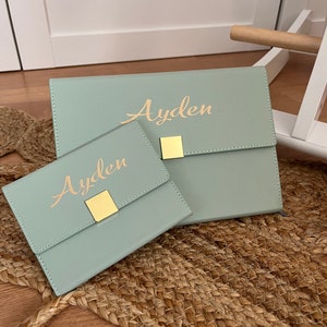 Peut inclure: Deux pochettes en similicuir vert clair avec des fermoirs dorés et le nom "Ayden" écrit en lettres dorées.