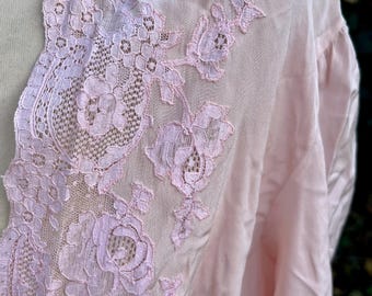 Op de afbeelding: Zachtroze badjas met ingewikkelde bloemenkanten afwerking. Het kant, in een lichtere roze tint, heeft gedetailleerde rozen- en bladerpatronen. De stof van de badjas heeft een zachte, vloeiende uitstraling.