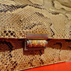 Op de afbeelding: Een vintage handtas met een slangenpatroon in tinten beige, bruin en zwart. De tas heeft een metalen sluiting en handvat. De textuur van de slangenhuid is zichtbaar, wat bijdraagt aan de unieke uitstraling.