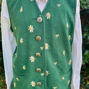 May include: A green vest with white daisy embroidery and gold-colored buttons. The vest has a V-neck and two front pockets. The daisies are scattered across the vest. The vest is worn over a white blouse.
