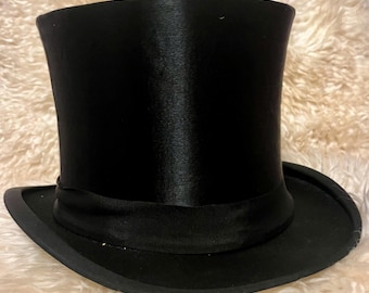May include: A classic black top hat with a tall, cylindrical crown and a wide, flat brim. The hat features a black grosgrain ribbon band around the base of the crown. The hat is made of a shiny, smooth material.
