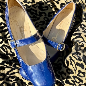 Può includere: Un paio di scarpe in vernice blu con cinturino con fibbia. Le scarpe hanno una punta quadrata e un tacco basso. Le scarpe sono del marchio "Tartan" e sono fatte in Inghilterra.