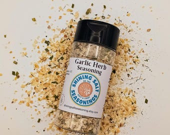 May include: A clear glass bottle of Garlic Herb Seasoning with a black cap, labeled "Shining Salt Seasonings." The bottle is surrounded by dried herbs and spices. The label features a sun graphic and the website shiningsaltseasoning.etsy.com.