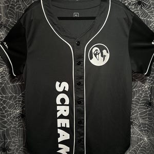 Peut inclure: Un maillot de baseball noir avec une bordure blanche et le mot "SCREAM" verticalement sur le devant. Comprend un graphisme circulaire blanc avec un motif de visage de fantôme. Le maillot a des manches noires et une fermeture boutonnée.