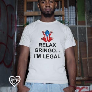 May include: White t-shirt with a red and blue frog wearing a Puerto Rican flag. The text on the shirt reads "Relax Gringo... I'm Legal."