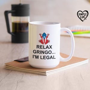 Puede incluir: Taza de café de cerámica blanca con una rana roja y azul que lleva una bandera puertorriqueña. La taza dice "Relax Gringo... I'm Legal".
