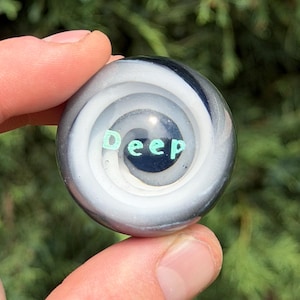May include: A small, round, glass marble featuring a swirling pattern of black and white colors. The center of the marble contains the word "Deep" written in light green, sans-serif capital letters, set against a dark, circular background.