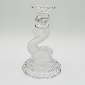 May include: A clear glass candlestick holder featuring a dolphin design. The candlestick has a round, patterned base, a dolphin-shaped stem, and a top designed to hold a candle. The glass is clear and reflects light.