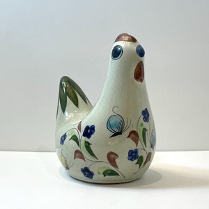 Puede incluir: Figura de gallina de cerámica con base color crema. La gallina está decorada con flores azules, hojas verdes y detalles marrones. La cresta de la gallina es marrón y sus ojos son azules. Es un objeto decorativo.