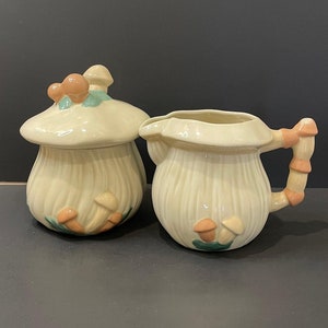 Op de afbeelding: Een crèmekleurige keramische suikerpot met deksel en een melkkan, beide in de vorm van paddenstoelen. De suikerpot heeft een paddenstoelvormig deksel met oranje en groene accenten. De melkkan heeft een paddenstoelvormig handvat.