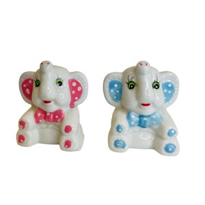 Puede incluir: Dos figuritas de elefante de cerámica, una con orejas de lunares rosas y una pajarita rosa, la otra con orejas de lunares azules y una pajarita azul.