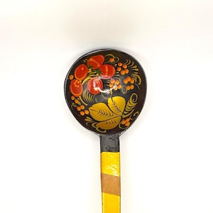 Op de afbeelding: Een houten lepel met een zwarte kom en een lange steel. De kom is versierd met rode bessen, oranje bessen en gele bladeren. De steel is geel, bruin en rood geverfd. Een decoratief keukengerei.