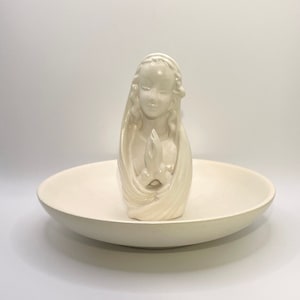 May include: A white ceramic bowl with a white ceramic figurine of a woman with her hands clasped in prayer. The figurine is in the center of the bowl.