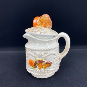 以下が含まれることがあります： キノコのデザインが施された白いセラミック製のクリーマー。クリーマーは、上にキノコが乗った蓋が付いています。クリーマーは、茶色とオレンジ色のキノコのデザインで飾られています。