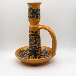 May include: A mustard yellow ceramic candle holder with a circular base, a central pillar, and a curved side handle. The surface is decorated with hand-painted dark blue floral patterns featuring small red centers on the petals and leaves.