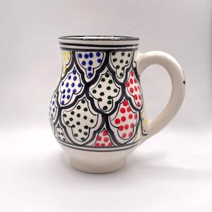 May include: A white ceramic mug with a black rim and handle. The mug is decorated with a geometric pattern of green, red, blue, and yellow dots.
