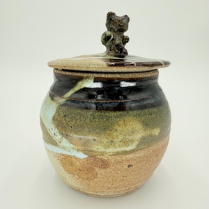 May include: A ceramic jar with a lid and a small bear figurine on top. The jar has a brown, green, and white glaze with a speckled finish.