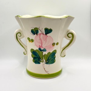May include: Cream-colored ceramic vase with a scalloped rim and two handles. The vase is decorated with a pink flower and green leaves. Green trim accents the rim and base. A decorative piece for the home.