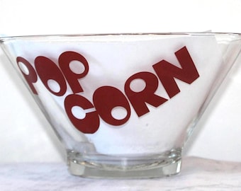 Op de afbeelding: Transparante glazen popcornschaal met rode letters. De kom heeft een breed, schuin ontwerp en een vierkante basis. Het woord "POP CORN" is vetgedrukt, zonder schreef op de zijkant van de kom gedrukt.