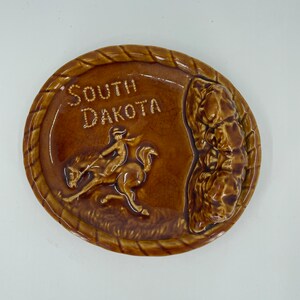 May include: Brown ceramic ashtray with a raised design of a cowboy riding a horse and the words "South Dakota".
