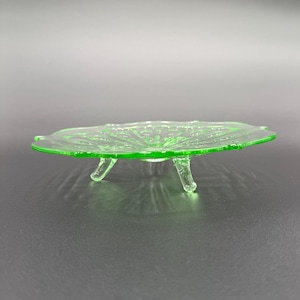 May include: A green glass pedestal dish with a scalloped edge and a textured surface. The dish has three legs and is perfect for serving snacks or desserts.