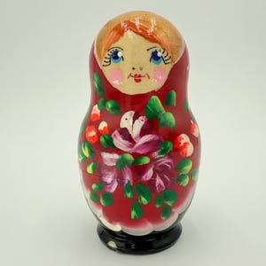 May include: A red Matryoshka doll with a painted floral design. The doll features a painted face with blue eyes, pink cheeks, and red lips. The floral design includes white, pink, and green accents. The base is black.