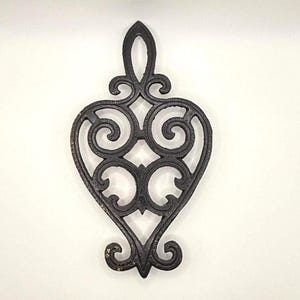 May include: Black cast iron trivet with a heart-shaped design. The trivet has detailed scrollwork and a loop for hanging. This decorative item is designed to protect surfaces from hot pots and pans.