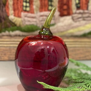 May include: A red glass apple with a green stem. The apple is sitting on a table with a green plant in the background.