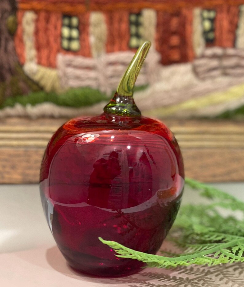 May include: A red glass apple with a green stem. The apple is sitting on a table with a green plant in the background.