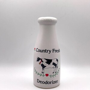 May include: A white ceramic bottle-shaped deodorizer featuring a black and white cow illustration with a red collar and bell. The text "Country Fresh" appears above the cow, and "Deodorizer" is printed below, framed by green leaves and a red heart.