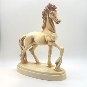 Op de afbeelding: Een keramieken paardenbeeldje in een staande pose, met een crèmekleurig lichaam en bruine manen en details. Het paard is gemonteerd op een crèmekleurige ovale basis. De sculptuur is ongeveer 25 cm hoog.