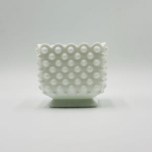 May include: A white hobnail glass bowl with a square base and scalloped rim. The bowl is covered in small, evenly spaced, raised dots. The hobnail design gives the bowl a textured appearance. The bowl is likely vintage and suitable for display or use.