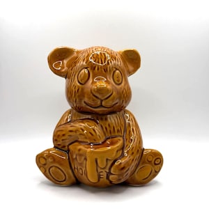 Puede incluir: Olla de miel de cerámica marrón con forma de oso, con un acabado brillante. El oso está sentado y sostiene una olla de miel con la palabra "HONEY" visible. La olla tiene un diseño detallado y es adecuada para la decoración de la cocina.
