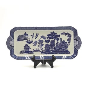 May include: A rectangular ceramic serving tray featuring a blue and white willow pattern. The design includes illustrations of pagodas, trees, and birds. The tray has a geometric border pattern and is supported by a black plastic display stand.