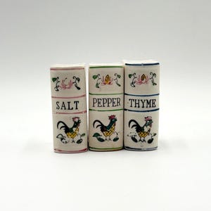 Może przedstawiać: Zestaw trzech ceramicznych solniczek i pieprzniczek z napisami "SALT", "PEPPER" i "THYME". Każda z nich ma ilustrację koguta i kwiatowe akcenty. Prostokątne pojemniki mają wygląd vintage.