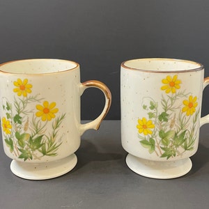 May include: Two white ceramic mugs with a brown rim and a footed base. The mugs feature a floral design with yellow flowers and green leaves on a speckled white background.