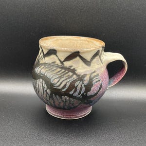 May include: A ceramic mug with a purple, black, and white design. The mug has a rounded body and a handle.