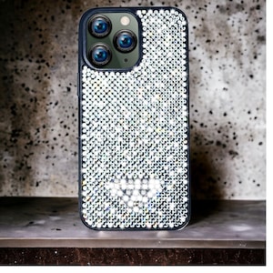 Puede incluir: Funda de teléfono adornada con cristales brillantes, con un borde azul oscuro. Presenta tres lentes de cámara. La funda está sobre una superficie oscura, con un fondo gris moteado.