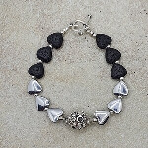 May include: A black lava rock and silver heart charm bracelet with a silver bead in the centre. The bead is decorated with small silver circles.