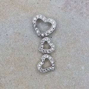 Puede incluir: Un colgante de plata con tres formas de corazón entrelazadas, cada una adornada con piedras brillantes transparentes.