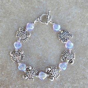 May include: A silver-toned bracelet featuring alternating sea turtle charms and iridescent, light purple beads. The bracelet has a toggle clasp and is displayed on a neutral-toned surface. The sea turtle charms are detailed with etched designs.