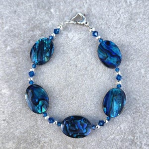 May include: A bracelet featuring oval-shaped, iridescent blue abalone shells. The shells are connected by small blue beads and silver-toned links. The bracelet has a heart-shaped clasp.