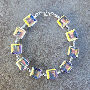 Op de afbeelding: Een armband met iriserende, vierkante kristallen aan een zilveren ketting. De kristallen tonen kleuren als geel, paars en blauw. De armband wordt gesloten met een zilveren sluiting.
